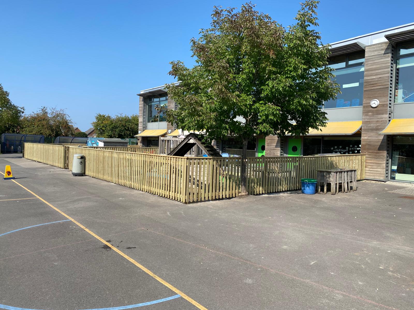 A school playground features a fenced area with a tree and a small wooden structure. The building in the background boasts large windows and yellow awnings. Painted lines for games mark the ground, surrounded by new gates installed by a commercial fencing contractor. A trash bin and cone sit on the pavement.