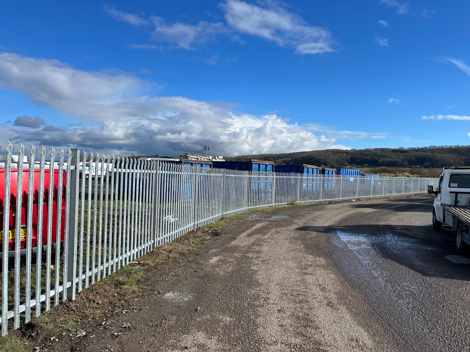 A rural road curves alongside a long metal fence installed by a commercial fencing contractor. The fence encloses an industrial area with blue and white containers. A white van is parked to the right, and a red vehicle is visible behind the fence under a partly cloudy, sunny sky.