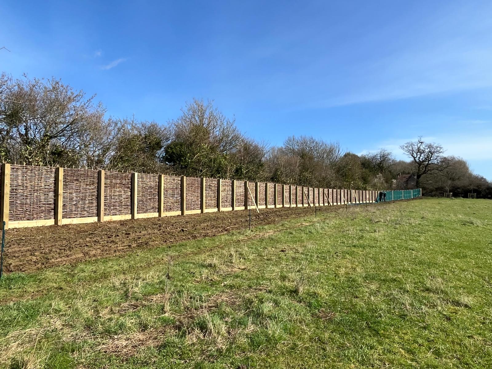 A long wooden fence stretches across a grassy field under a clear blue sky. Trees and shrubs line the bespoke railings, marking the boundary between the field and the forested area in the background.