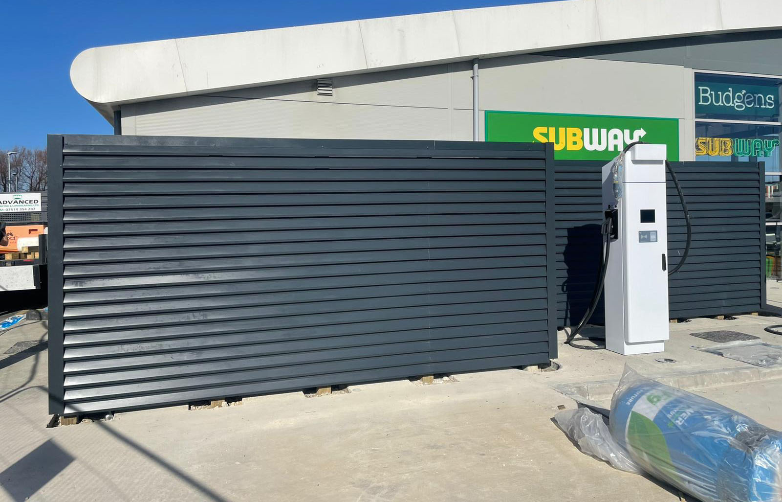 A large metal enclosure, crafted by a commercial fencing contractor, stands at a petrol station with a charging station to the right. The building behind displays signs for "Budgens" and "Subway," all under a clear blue sky.