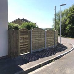 A row of wooden fences with latticed panels line a sidewalk on a sunny day, reminiscent of the artistry by a skilled fencing installation team. As the street curves alongside, with a lamppost and greenery in the distance, shadows from the fence are elegantly cast on the pavement.