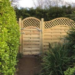 A wooden garden gate with an arched lattice top stands gracefully among lush hedges and plants. A narrow stone path, crafted with hard landscaping expertise, leads to this bespoke entrance. The overcast sky adds a serene atmosphere to the garden setting.