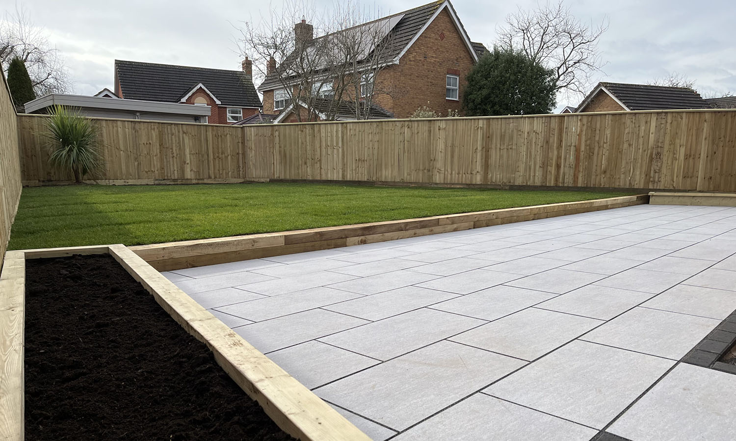 A backyard features a freshly laid tiled patio, a small raised garden bed with dark soil, and a lush green lawn. Enclosed by a sturdy wooden fence, expertly installed by a commercial fencing contractor, two brick houses stand visible in the background.