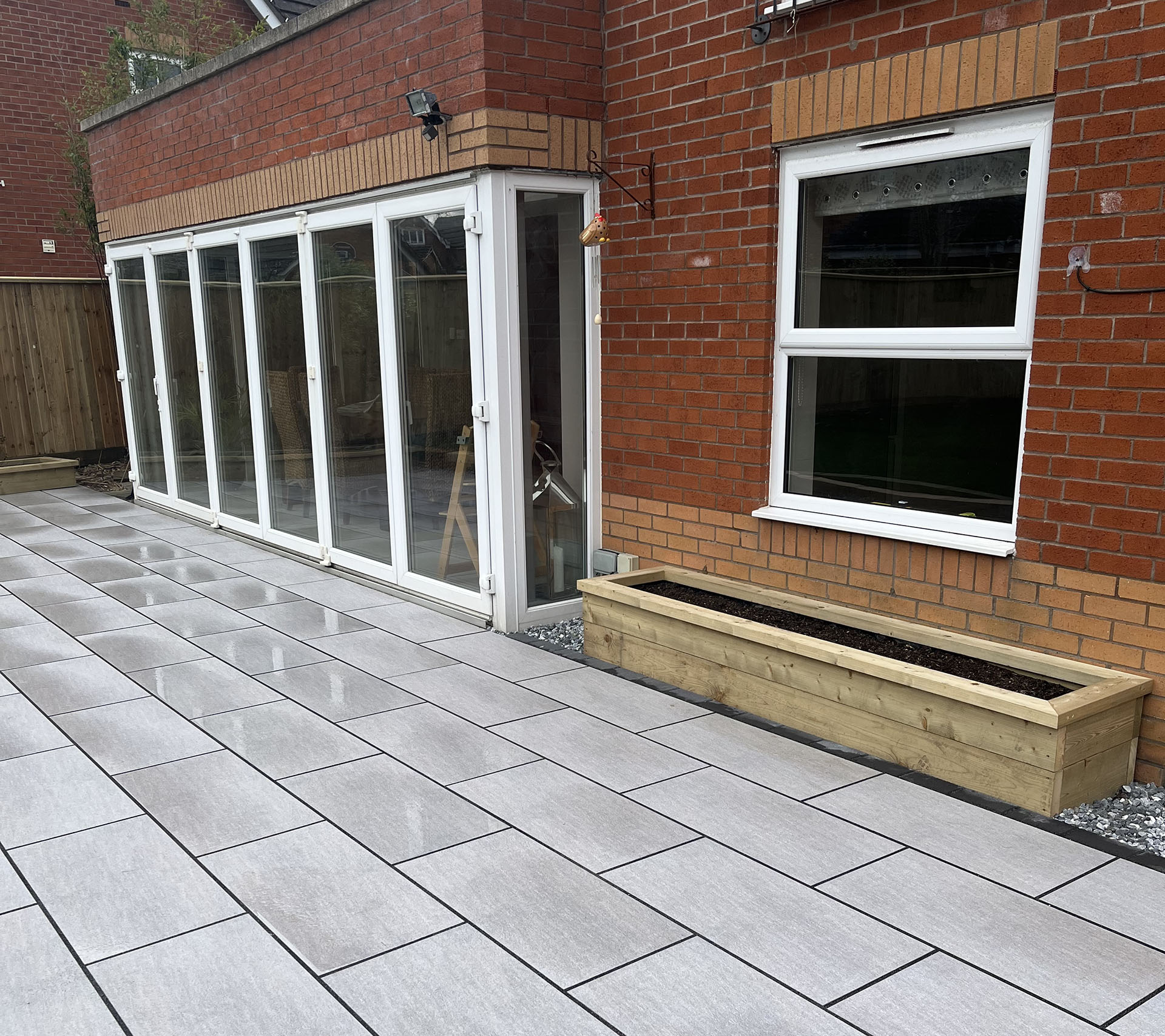 A modern patio with large rectangular gray tiles is shown. A red brick house with glass doors opens to the patio. Bespoke railings elegantly frame the space, while a wooden planter box is positioned beside a window on the house's side. Tight gravel borders the tiles.