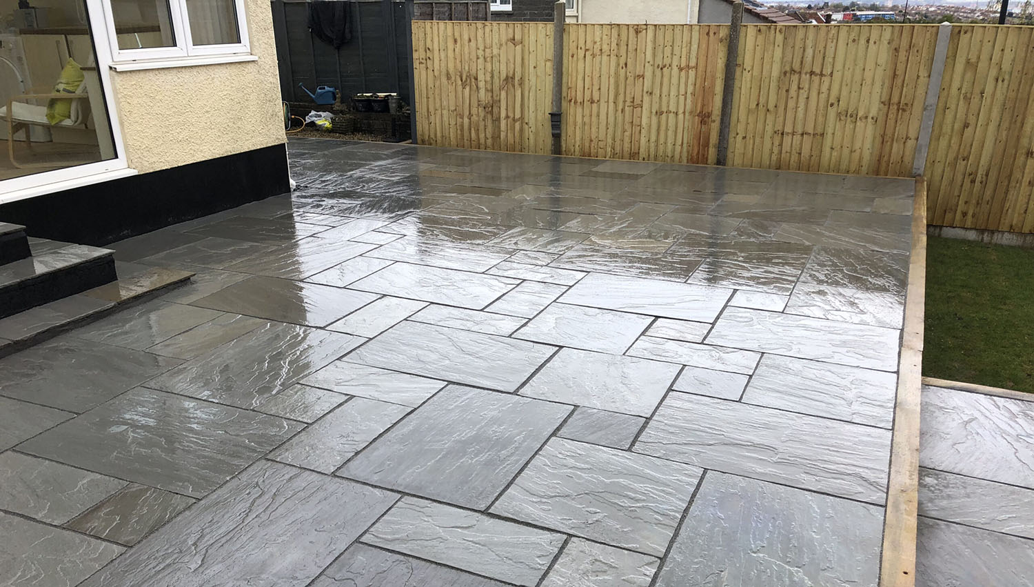 A backyard featuring a newly laid patio with large, wet grey tiles. The patio, bordered by bespoke railings, leads up to a light-colored house with a window and steps. A small patch of grass is visible on the right.