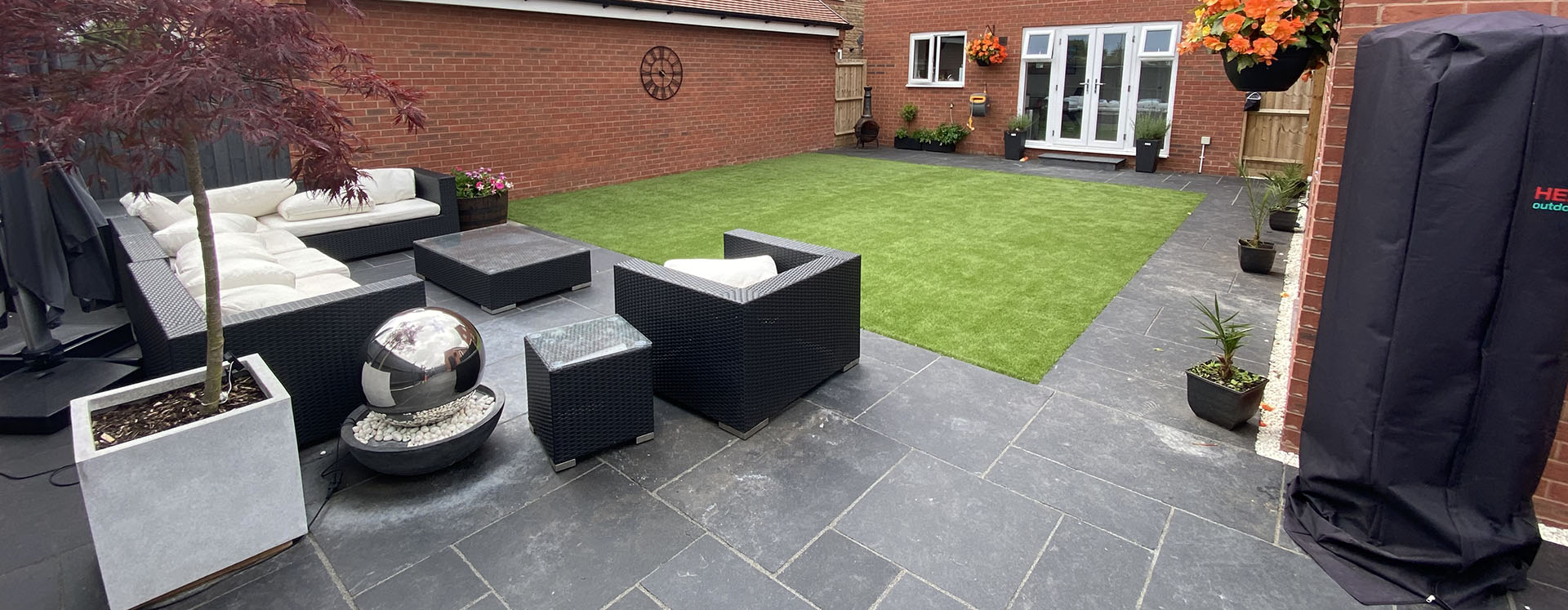 The backyard features artificial grass and sleek black patio tiles alongside modern outdoor furniture, including a sofa and chairs. Bespoke railings add a touch of elegance, while a small potted tree stands in the foreground and vibrant flowers adorn the brick wall. Large windows overlook this stylish haven.