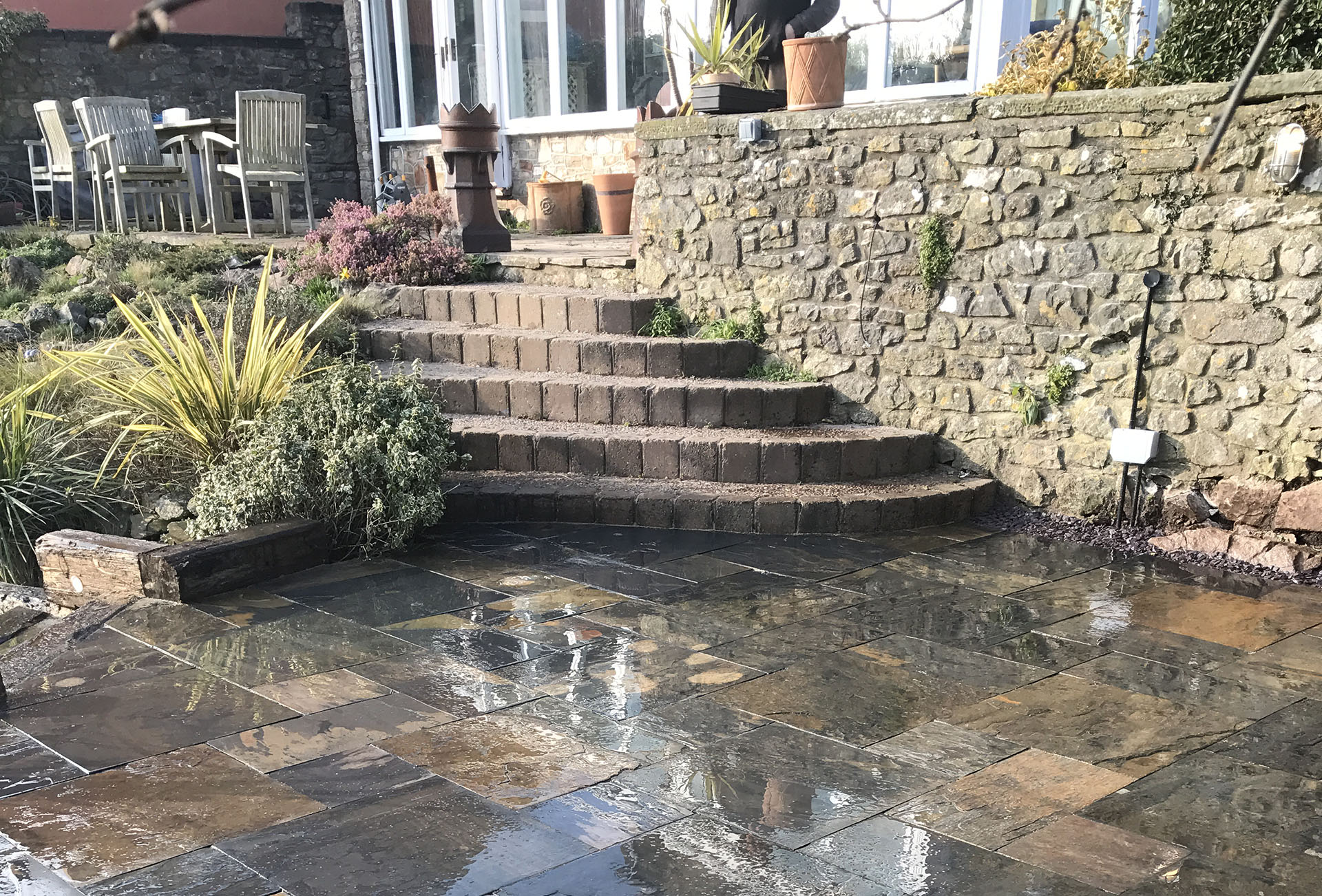 A sunlit patio with wet stone tiles leads to curved stone steps ascending to a garden area, enhanced by bespoke railings for safety. The terrace above boasts wooden chairs around a table and various potted plants. A stone wall borders the garden, featuring light fixtures and lush greenery.