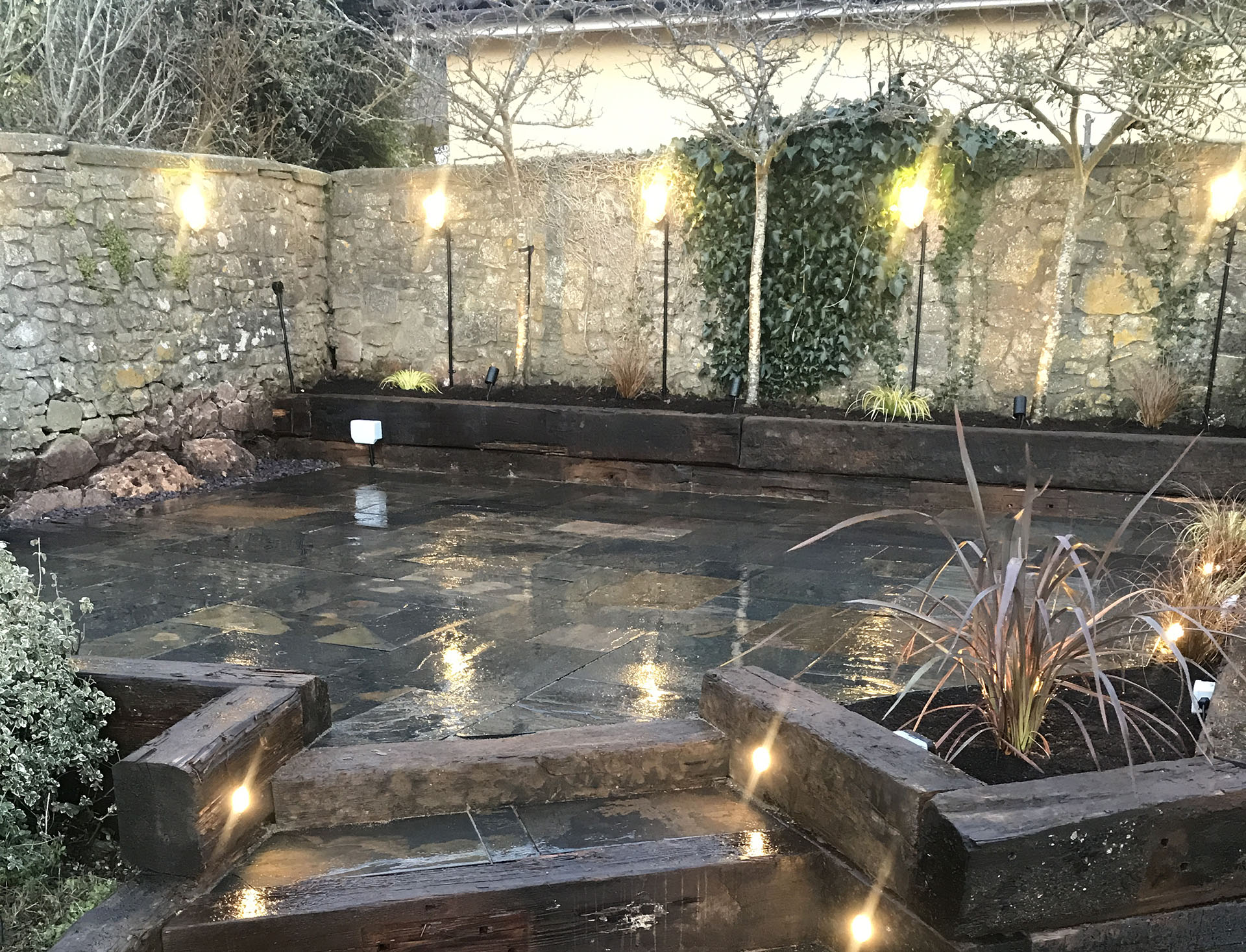 Wet stone patio with ambient lighting, surrounded by a stone wall with ivy and small trees. Raised wooden planters hold ornamental grasses, complementing the cozy atmosphere. Recently added hard landscaping enhances the design, ensuring a seamless blend of nature and function.