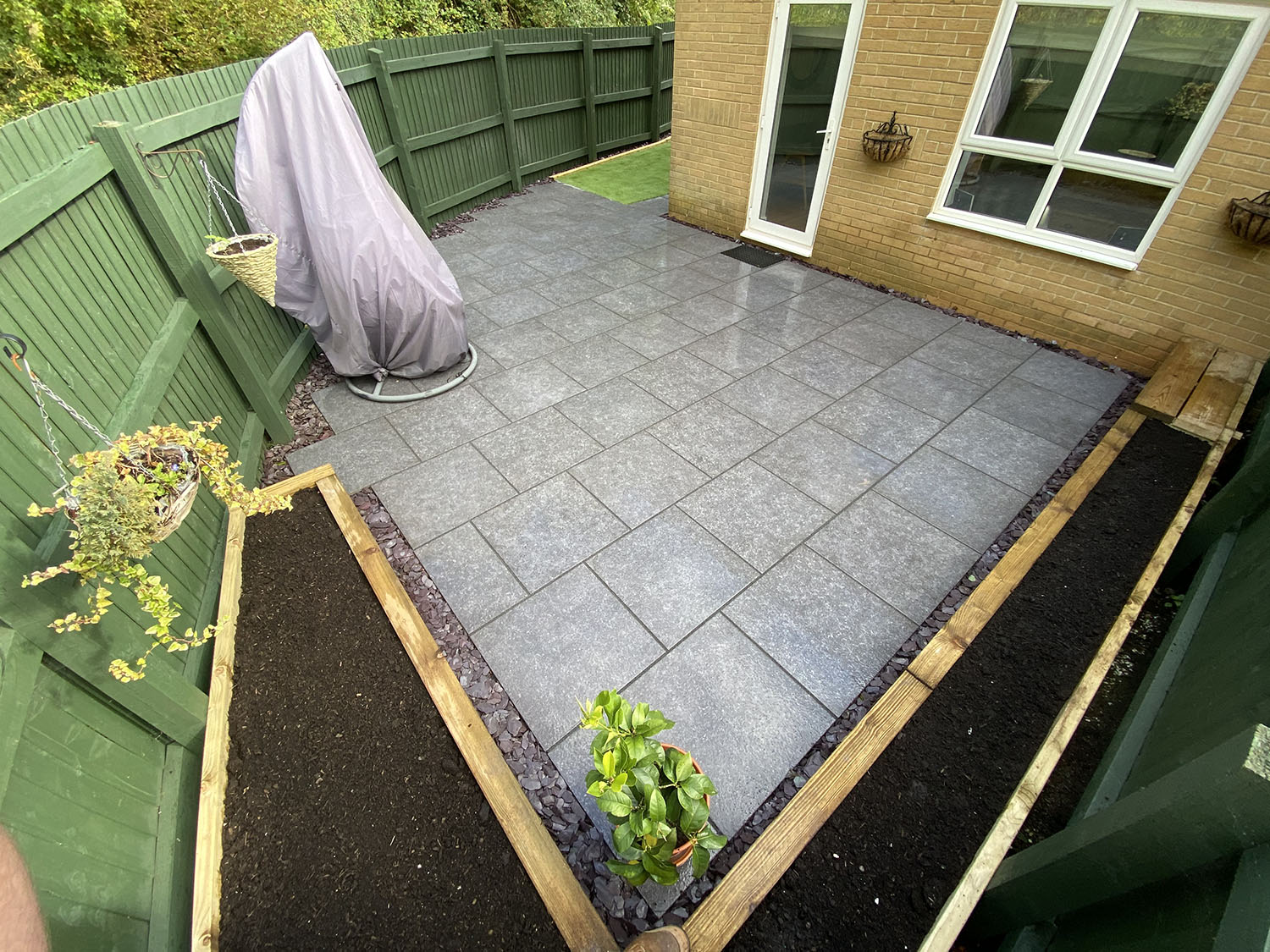A small backyard patio with gray tiles, bordered by bespoke railings. A covered swing chair rests in the corner. Raised garden beds line the sides with rich soil and a few plants. A door and windows punctuate the adjoining brick wall, enhancing this charming outdoor retreat.