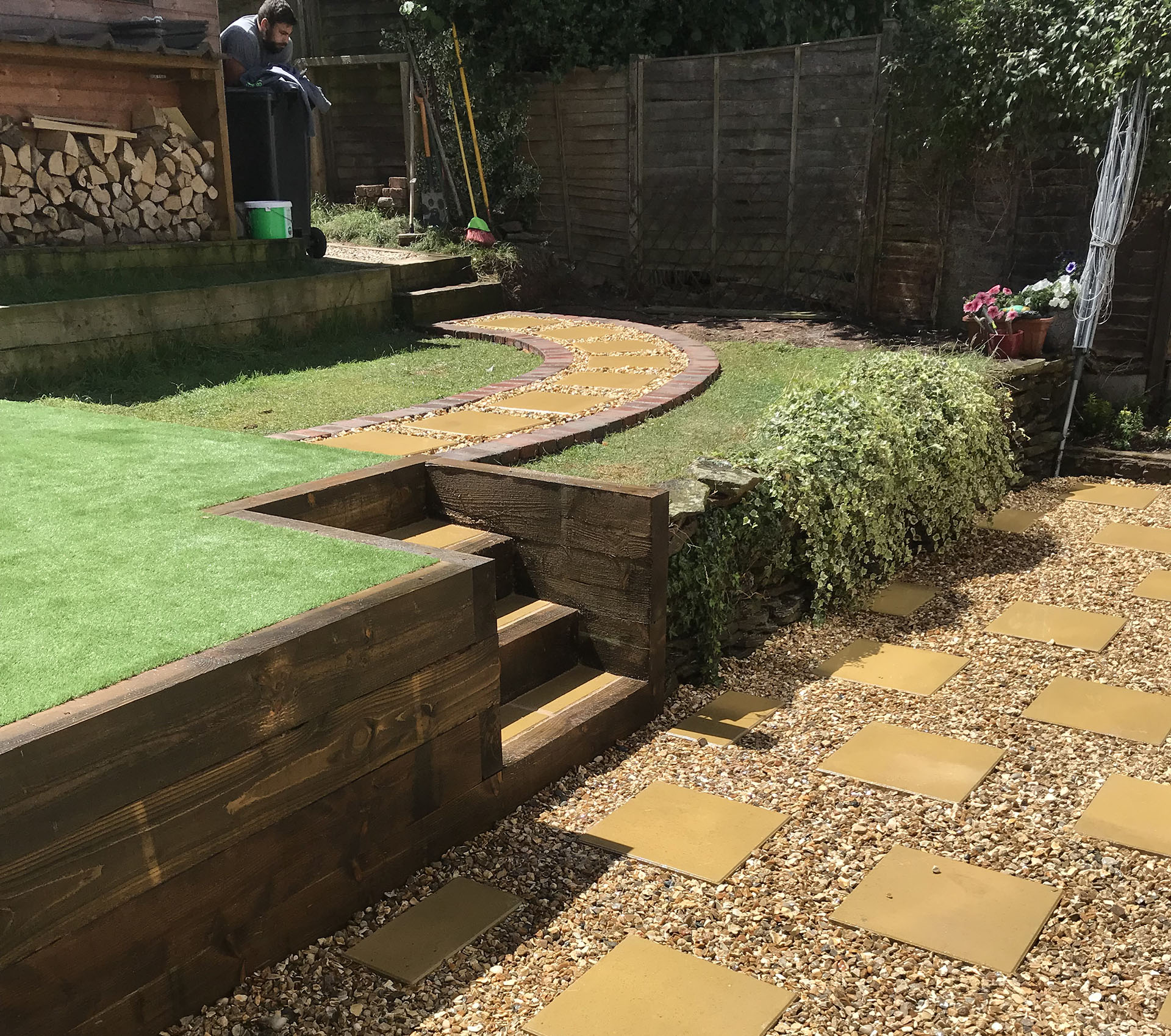 Backyard with a layered design featuring expert hard landscaping. The lower level boasts yellow square pavers and gravel, leading to two wooden steps. A curved stone path ascends to a lawn area, bordered by professional fencing installation, stacked firewood, and lush plants on the right.