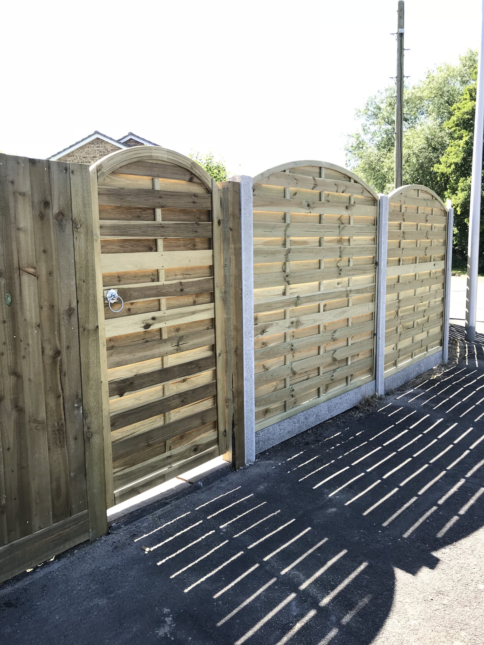 Wooden fence with three curved-top panels and a matching gate in a sunny outdoor setting, showcasing expert fencing installation. Shadows from the slats create striped patterns on the ground. A portion of a building and trees are visible in the background.