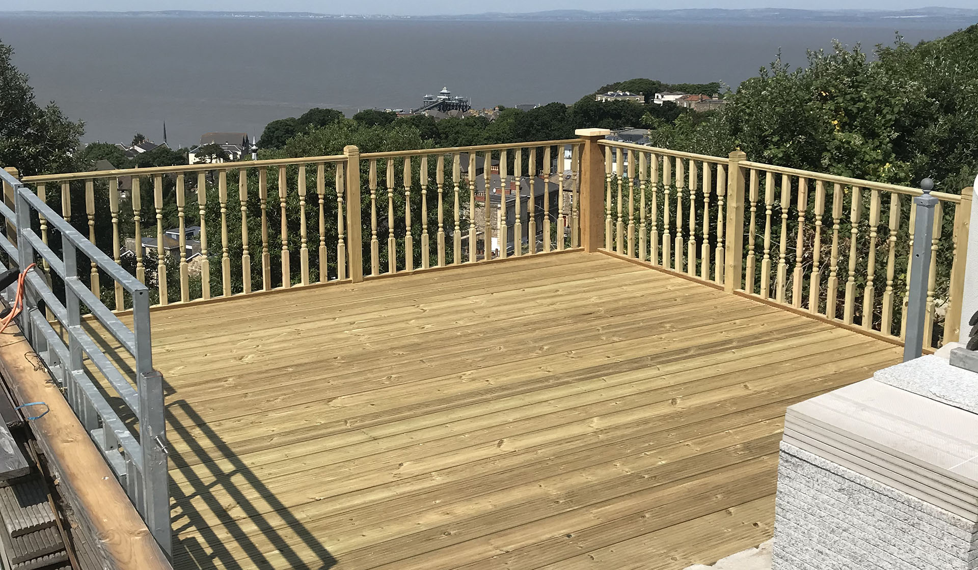 A wooden deck with new gates and railing overlooks a scenic view of the sea and distant hills. Elevated and surrounded by trees, it sits beneath a clear blue sky, with a few houses visible below the deck.