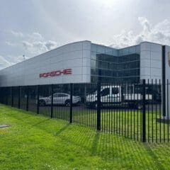 A modern Porsche dealership with a sleek glass and metal facade stands proudly behind bespoke railings. The Porsche logo is displayed on the building, while a white car and truck are parked in front. The sky is partly cloudy, adding to the contemporary charm of the scene.