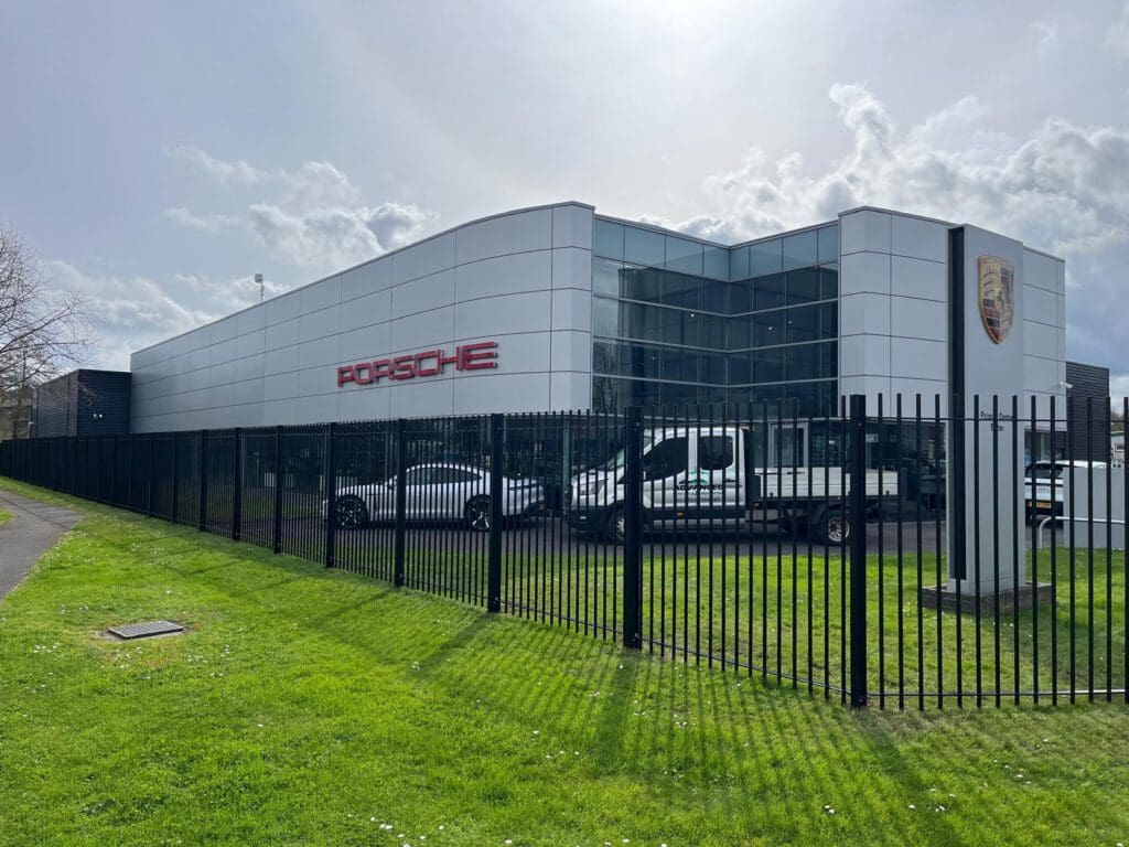 A modern Porsche dealership building with large glass windows and a white exterior. Several cars are parked in front, visible through bespoke railings. The Porsche logo and name are prominently displayed on the facade.