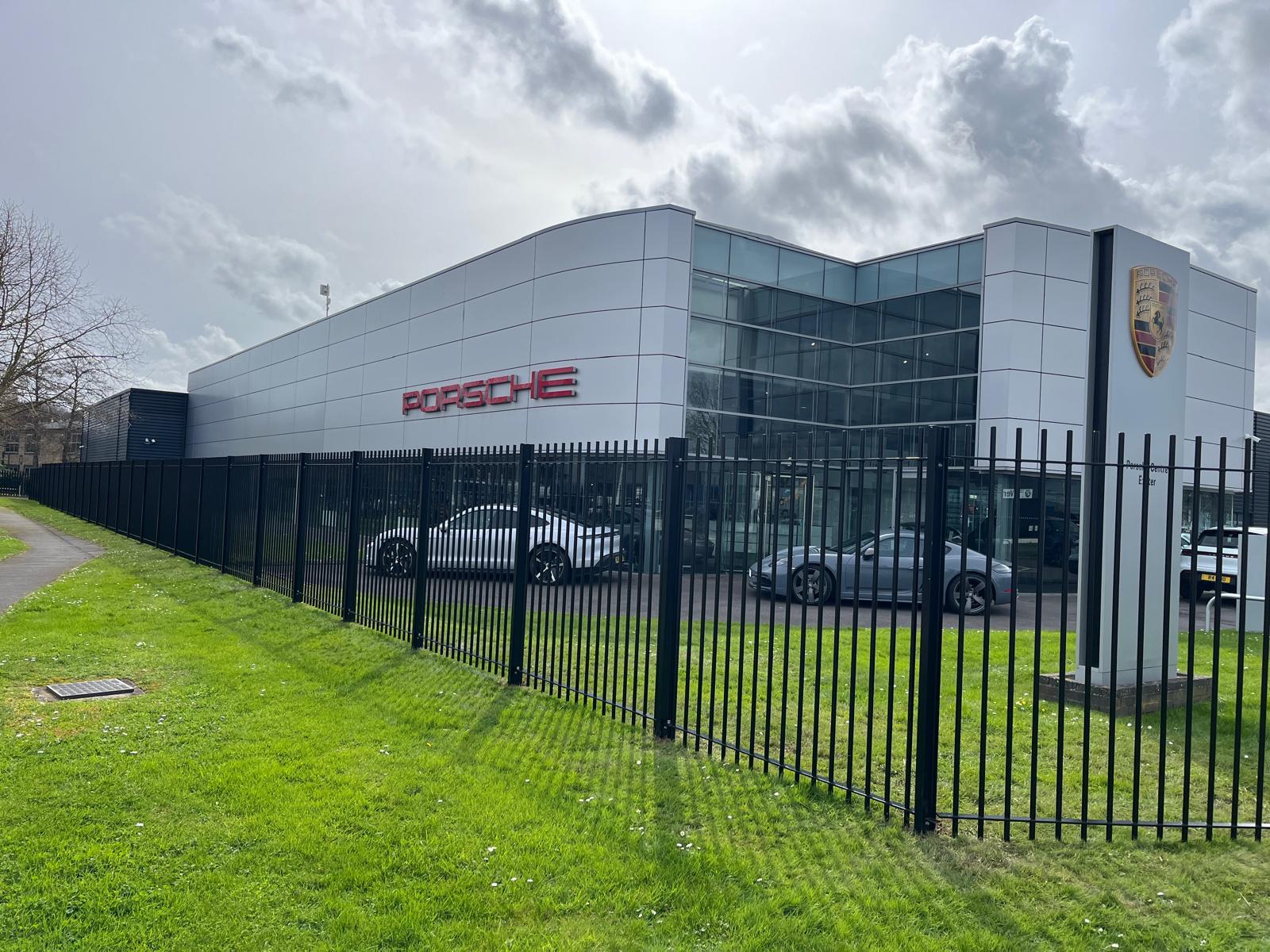 A modern Porsche dealership with a glass-front exterior and silver facade, where the logo and name are prominently displayed. Several cars are visible through the windows. The building features bespoke railings along a black metal fence, complemented by a grassy area in the foreground.