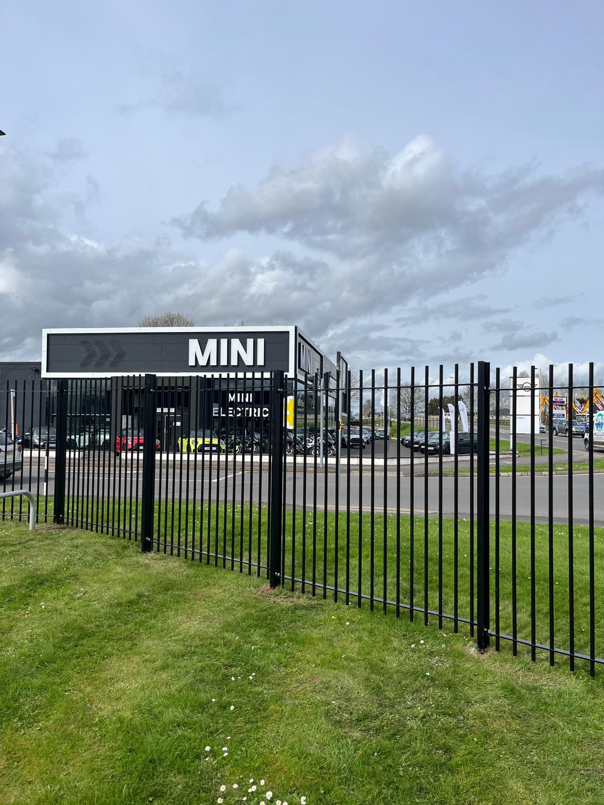 A sleek black metal fence, possibly installed by a commercial fencing contractor, frames the bustling car dealership with "MINI" signage. Adjacent to a busy road and under a partly cloudy sky, cars populate the lot against a backdrop of lush green grass.