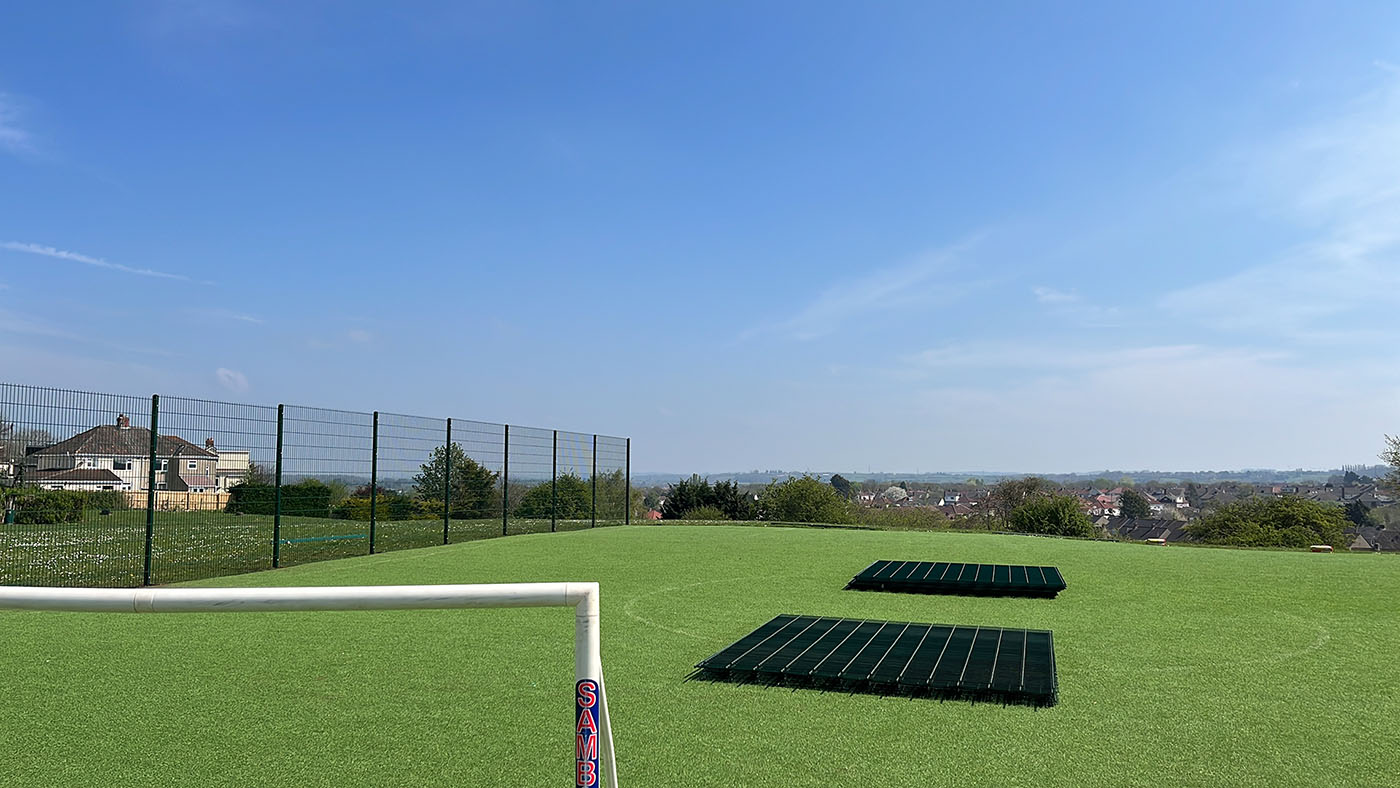 A grassy sports field features a white goalpost on the left and two rectangular sets of bleachers. Enclosed by bespoke railings, houses and trees grace the backdrop under a clear blue sky.