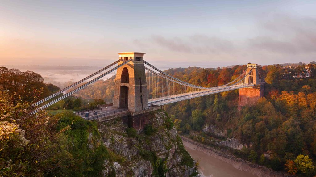The Clifton Suspension Bridge stretches across the Avon Gorge, surrounded by autumn trees and bathed in soft morning light with mist in the distance.