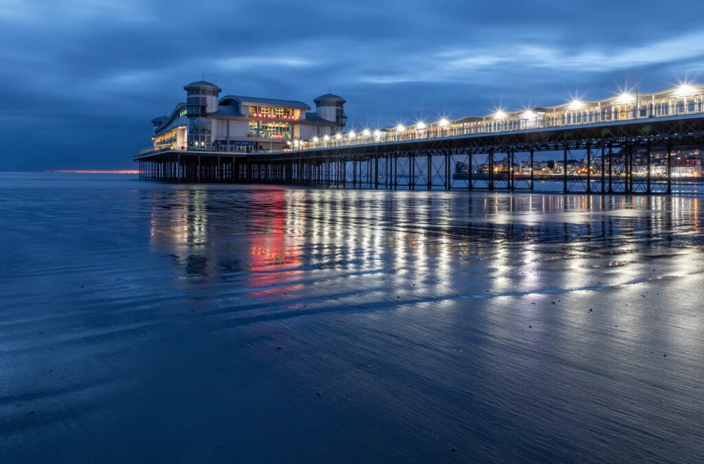 A brightly lit pier stretches over calm water at dusk, with buildings on the pier and reflections of lights shimmering on the wet sand and sea under a moody, cloudy blue sky.