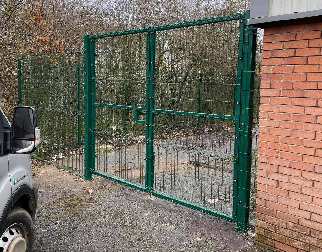 A green metal security gate next to a brick building, partially open, with trees and scattered litter visible in the background. The front of a white van is parked on the left side of the image.